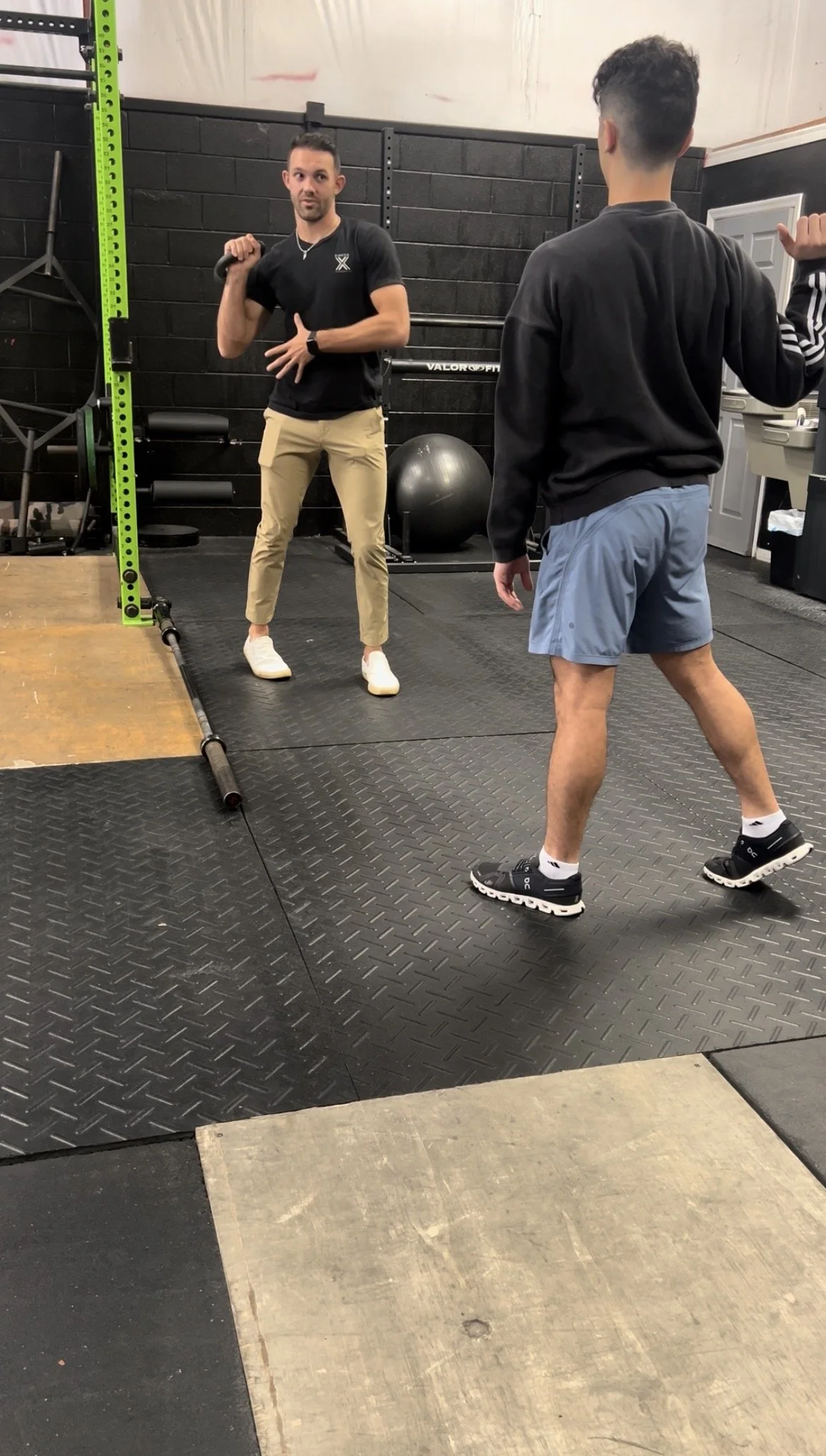 Two men in a gym, one holding a kettlebell on his shoulder and the other giving instructions. The gym has black walls, exercise equipment, a medicine ball, and a lifting platform.