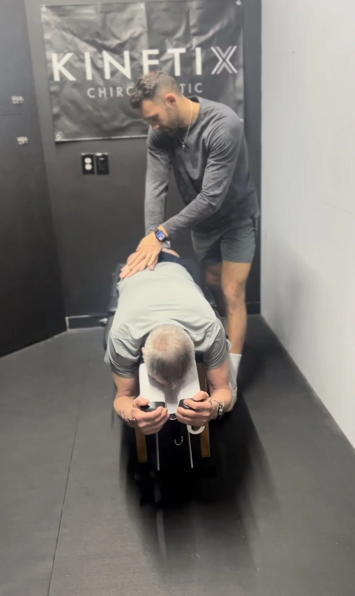 A chiropractor performing an adjustment on a patient lying face down on an adjustment table, with another person standing beside them in a chiropractic clinic.