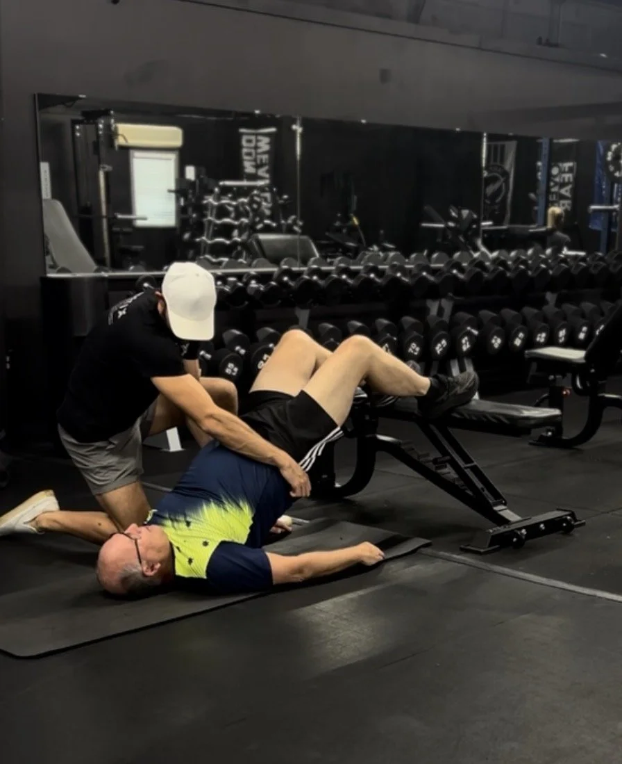 A man lying on a gym mat performing a Pilates exercise, assisted by another man in a gym setting with dumbbells and workout equipment in the background.