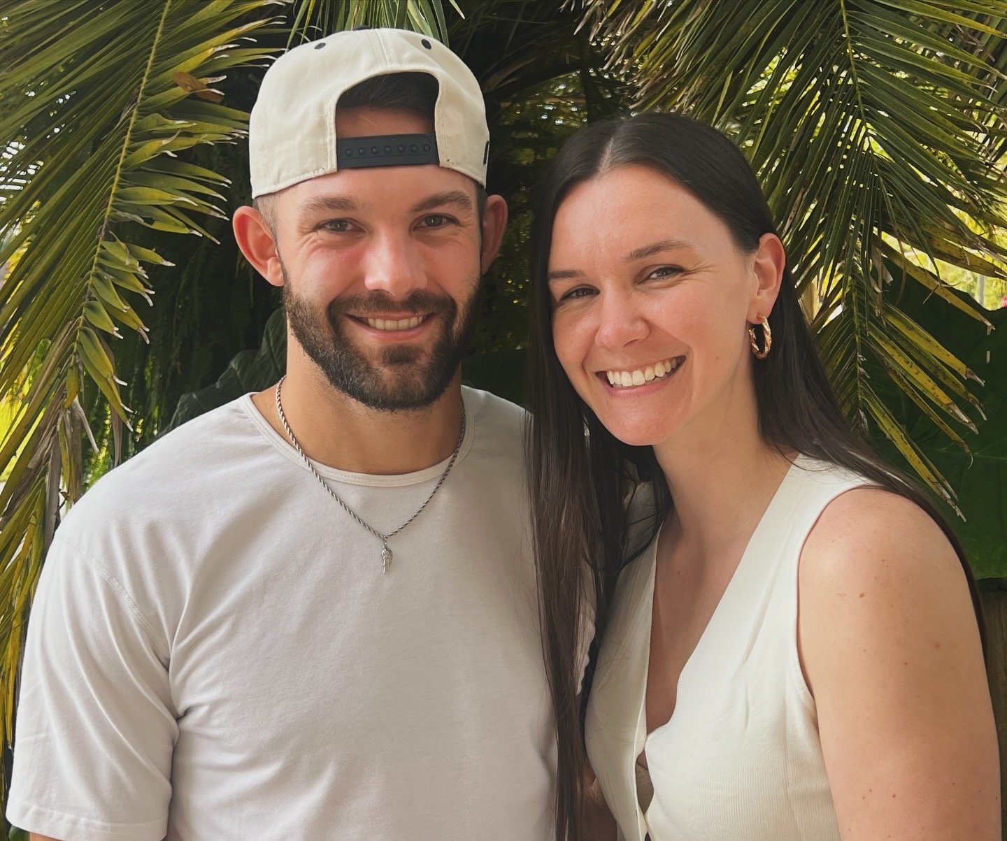 A man and woman smiling outdoors with tropical palm trees in the background.