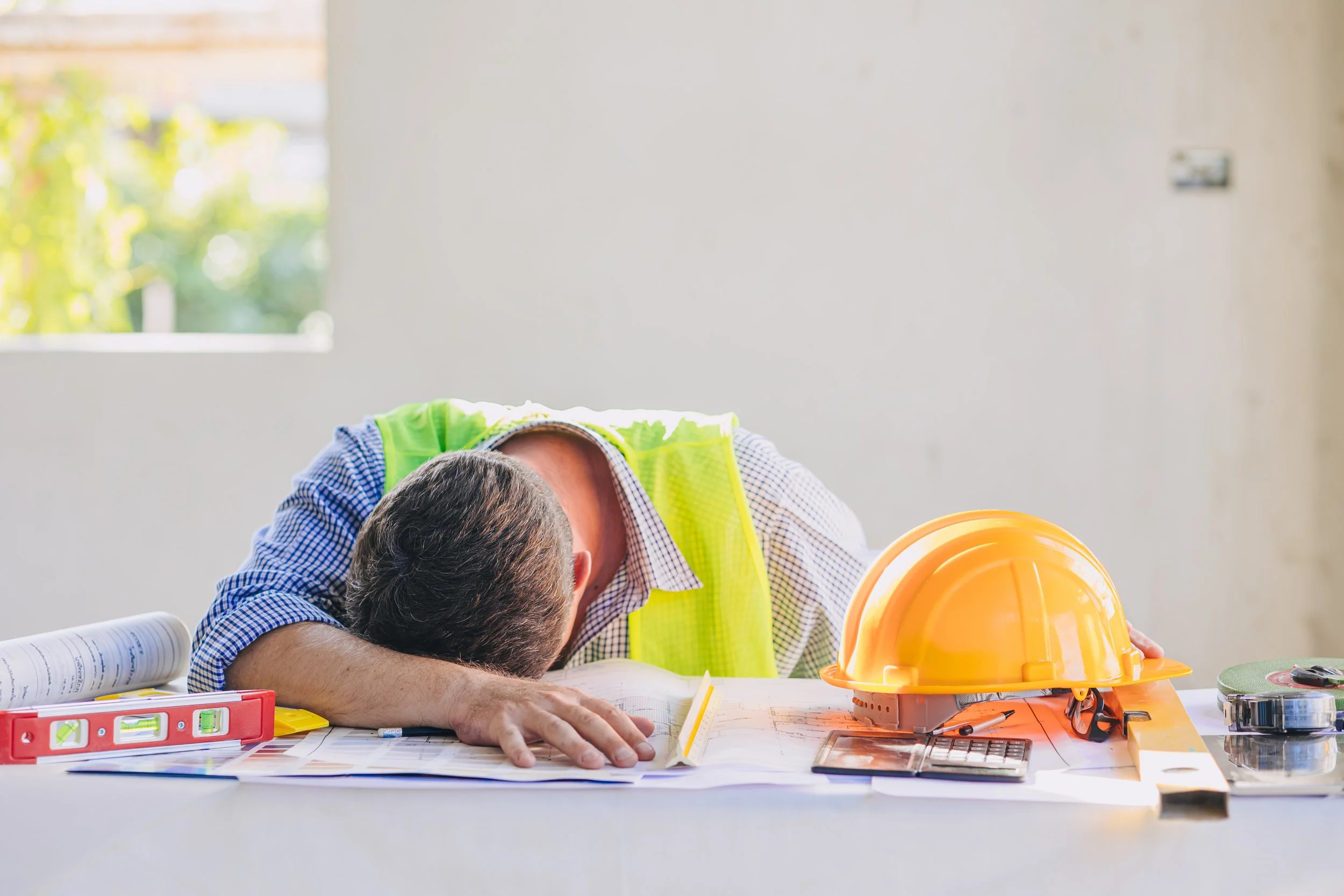 A construction worker wearing a safety vest resting his head on a table with construction tools, blueprints, a hard hat, and a spirit level.