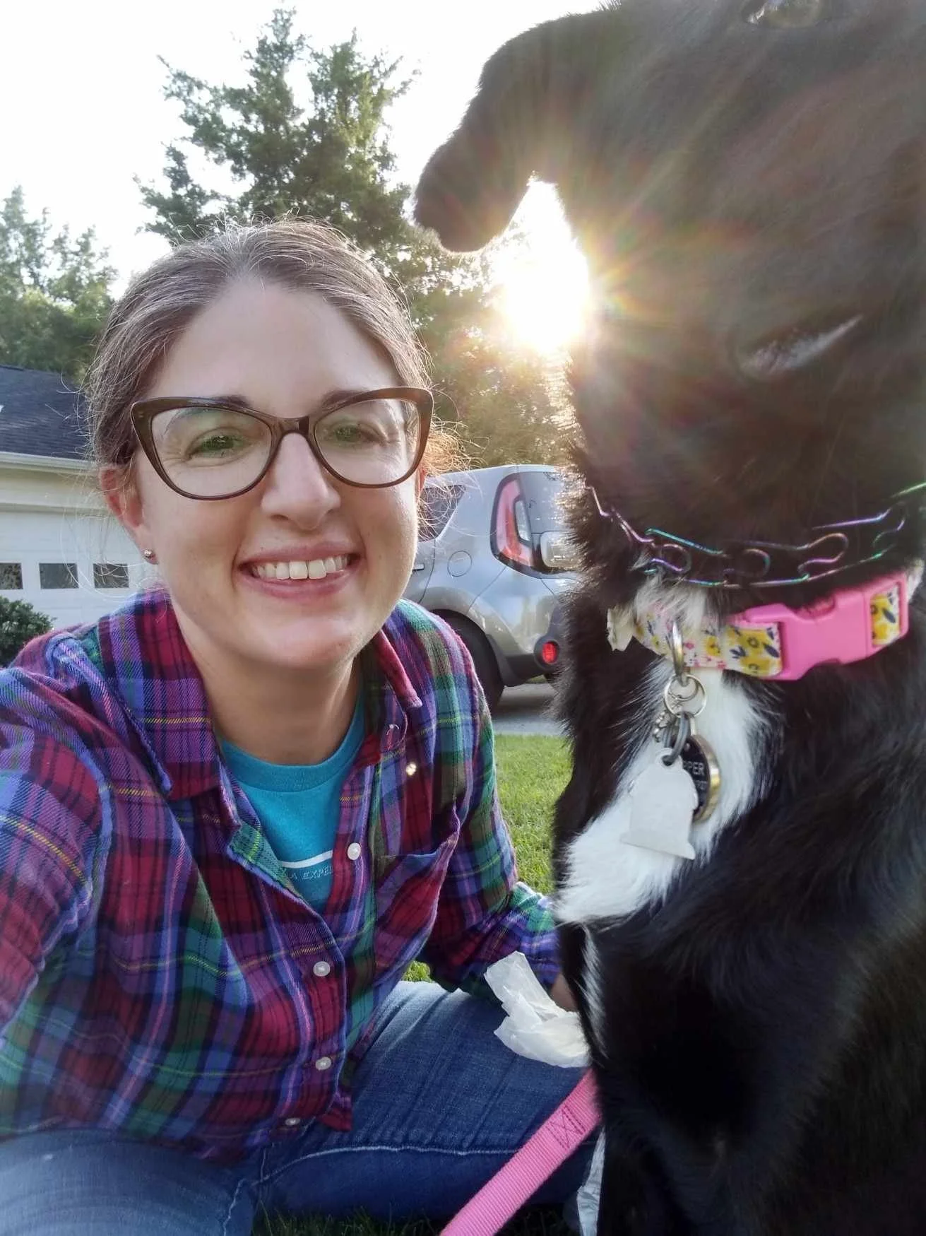 A woman with glasses, smiling, posing outdoors with a black and white dog wearing a pink collar and a yellow bow. The woman is wearing a plaid shirt and a blue t-shirt, with sunlight shining behind her, and a house and car in the background.