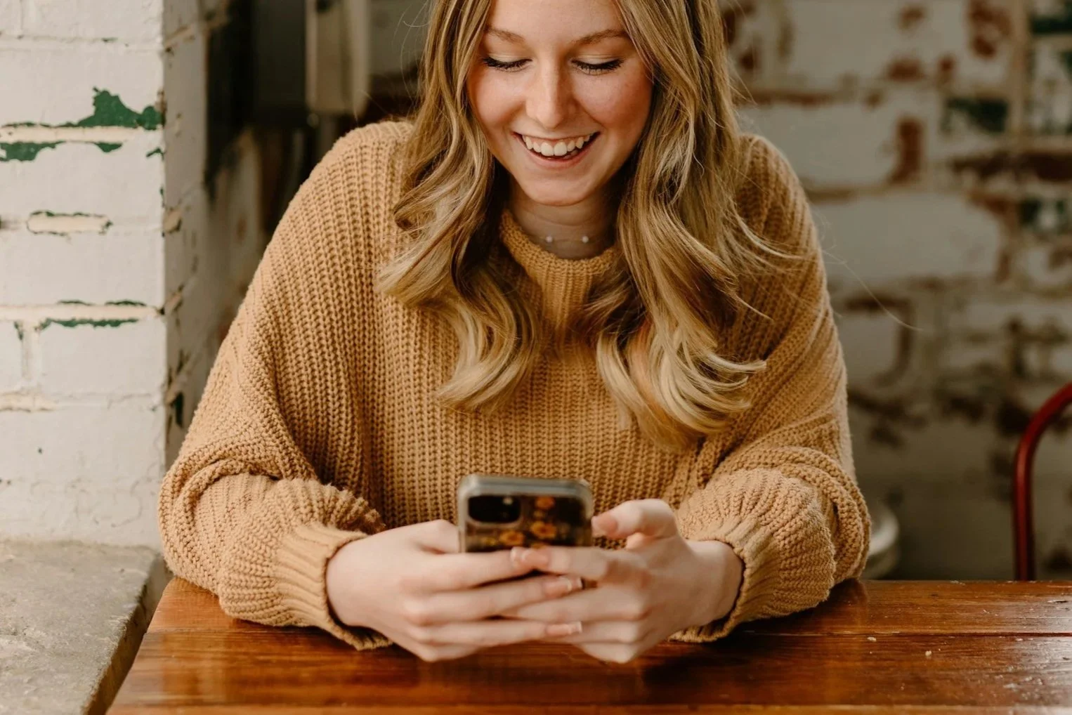 A woman with wavy blonde hair smiling while looking at her phone, sitting at a wooden table in a cafe.