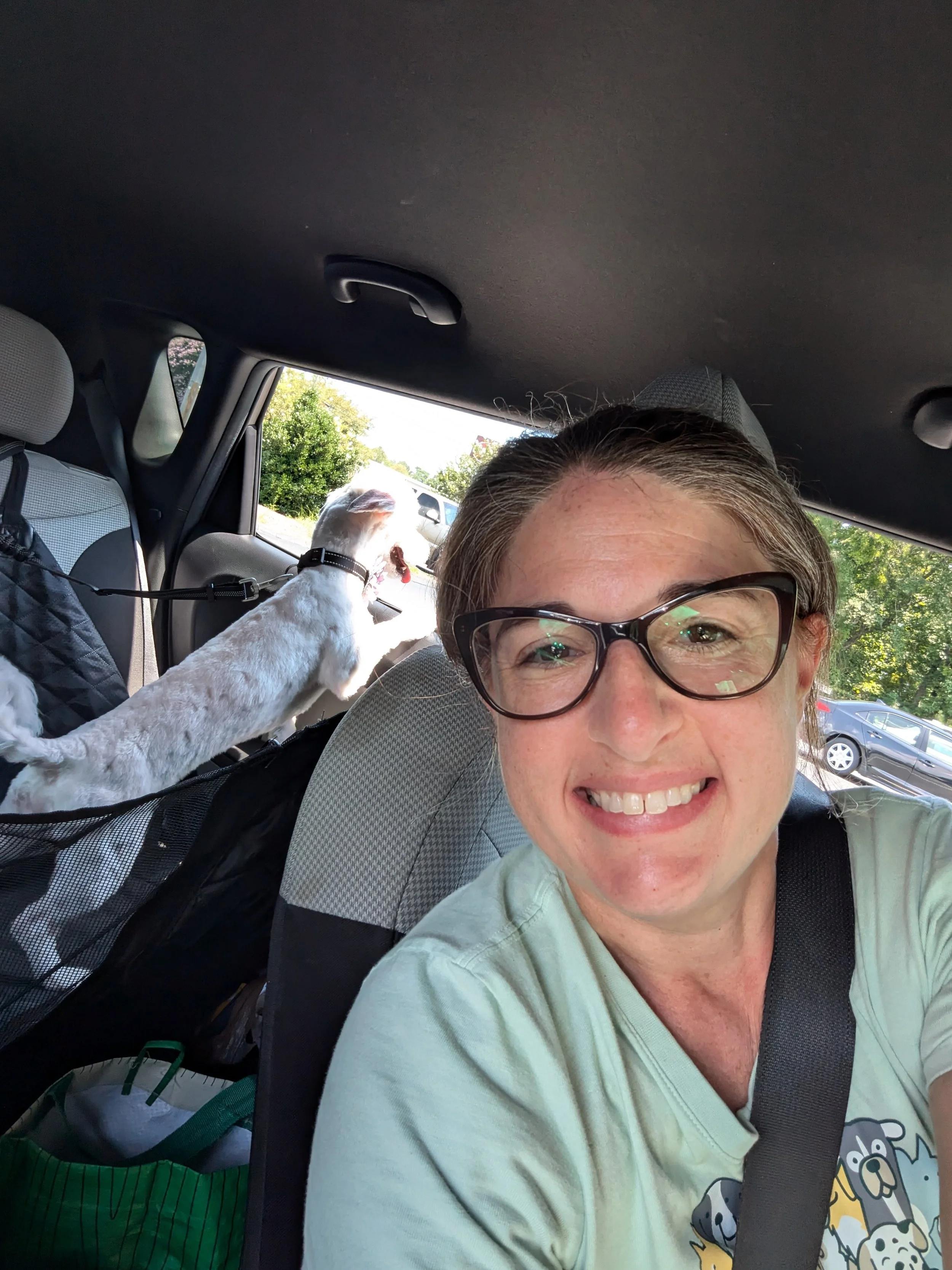 Young woman smiles in for ground, wearing glasses and with hair pulled back as a white small pup has his paws up on the door in the back seat showing him riding in a car and enjoying it, and smiling with his head out the window.