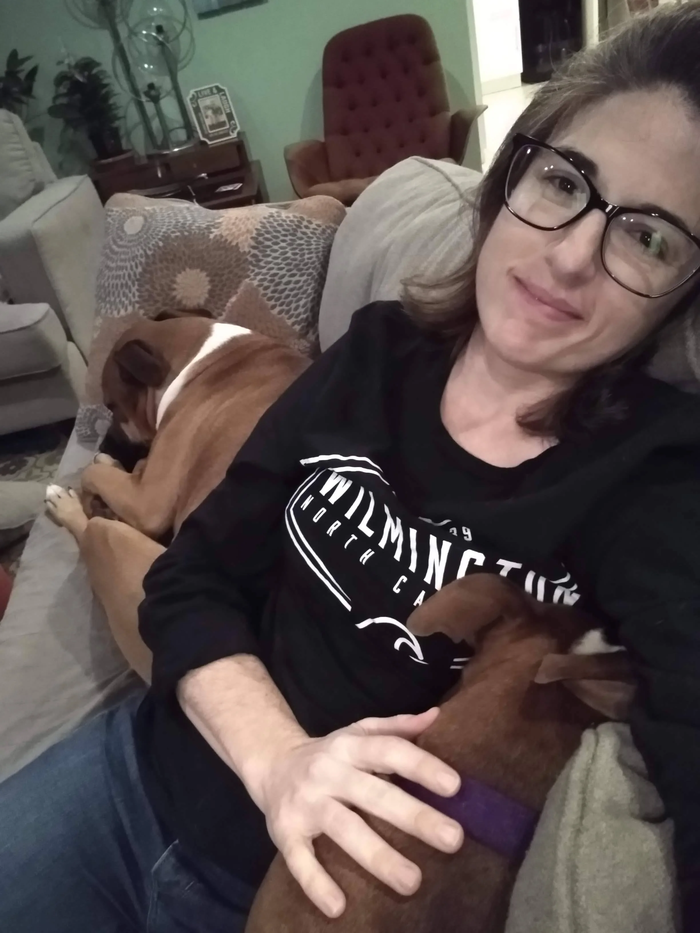 A woman wearing glasses and a black shirt is sitting indoors on a sofa, holding two dogs, a brown one and a smaller dark-colored one, close to her. There is a brown armchair, a small table with a decorative object, and some plants visible in the background.