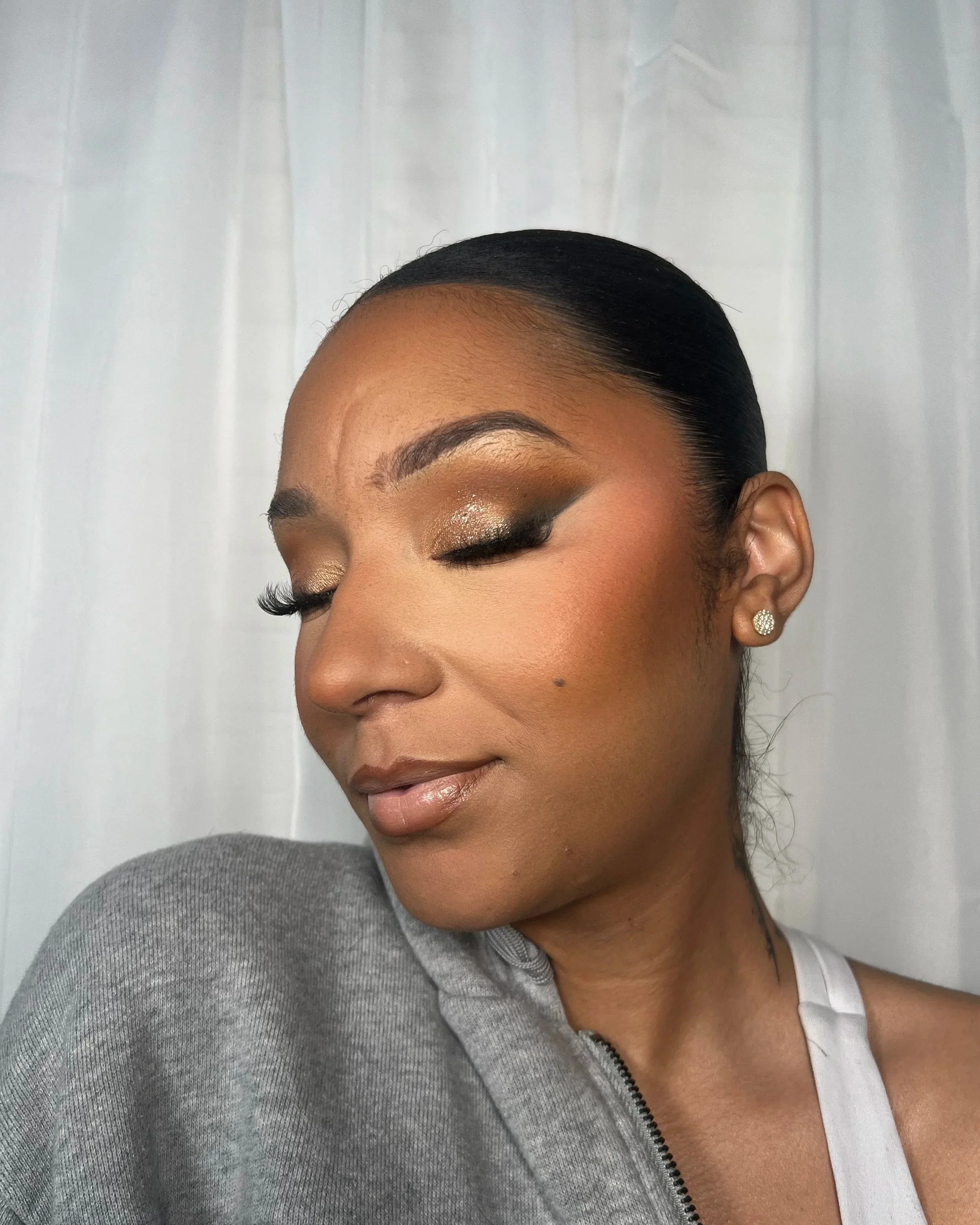 A woman with her eyes closed, showcasing her makeup. She has dark hair pulled back, gold eyeshadow, black eyeliner, and false eyelashes. She is wearing pearl earrings, a white top, and a gray jacket over her shoulder, with a white curtain background.