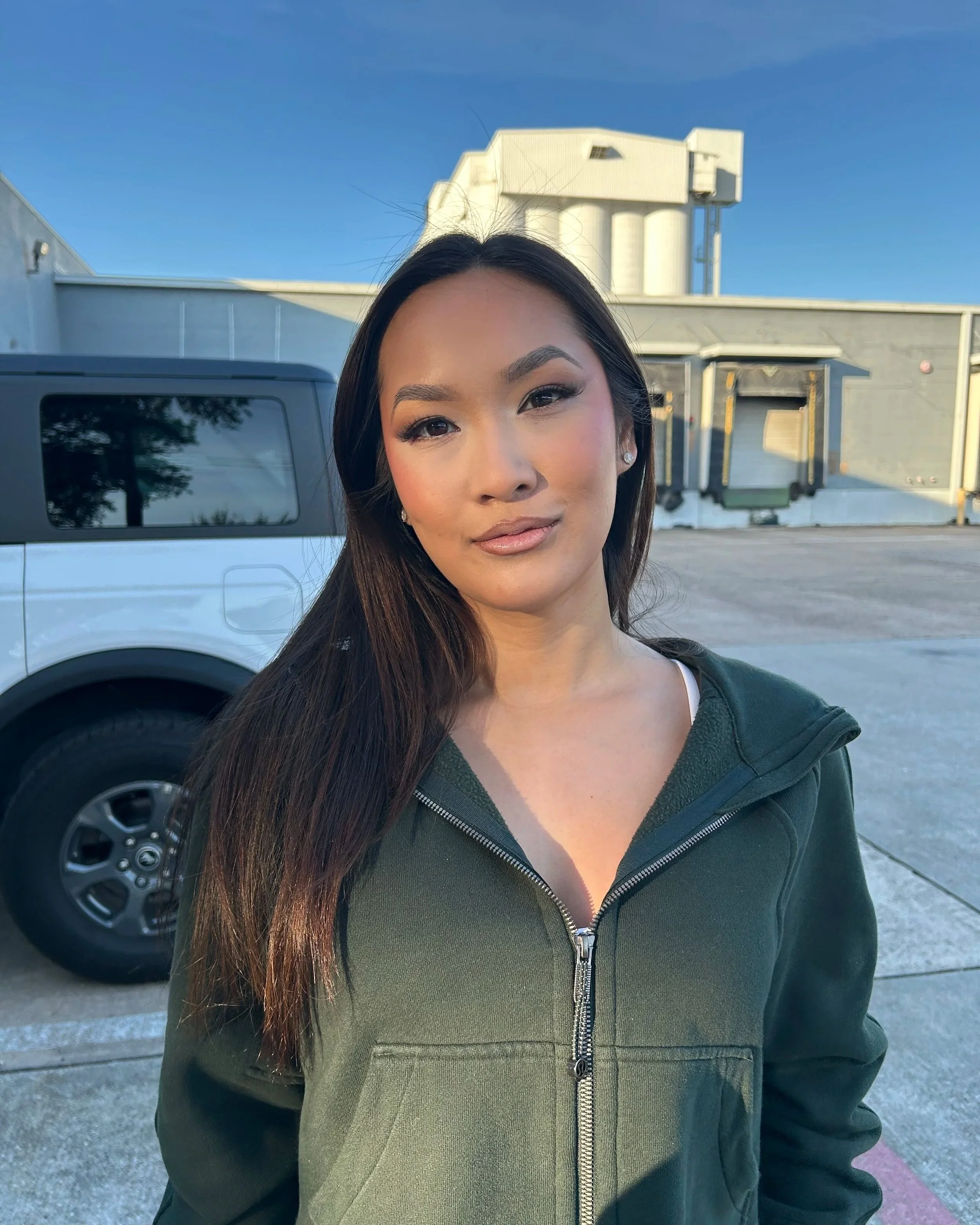 A young woman with long dark hair and makeup, wearing a dark green zip-up jacket, standing outdoors in front of a parked vehicle and a building with a rooftop structure, under a clear blue sky.