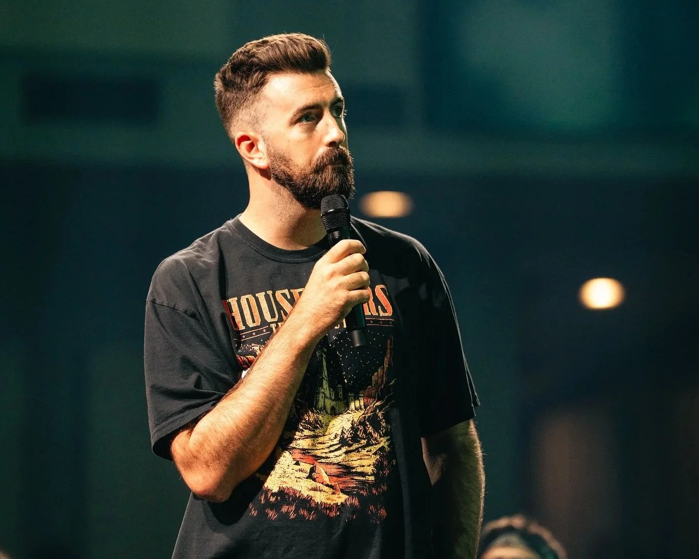 A man with a beard and styled hair holding a microphone, standing on a dark stage.