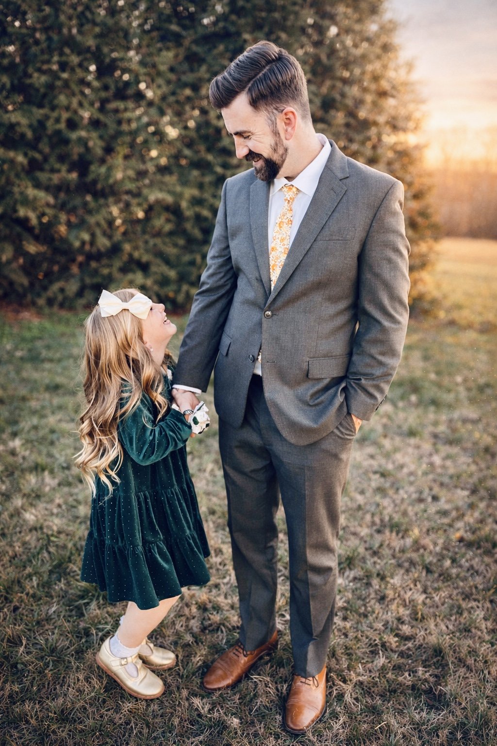 A man in a gray suit and a girl with long blonde hair wearing a dark green dress and white shoes standing outdoors during sunset, smiling and looking at each other.