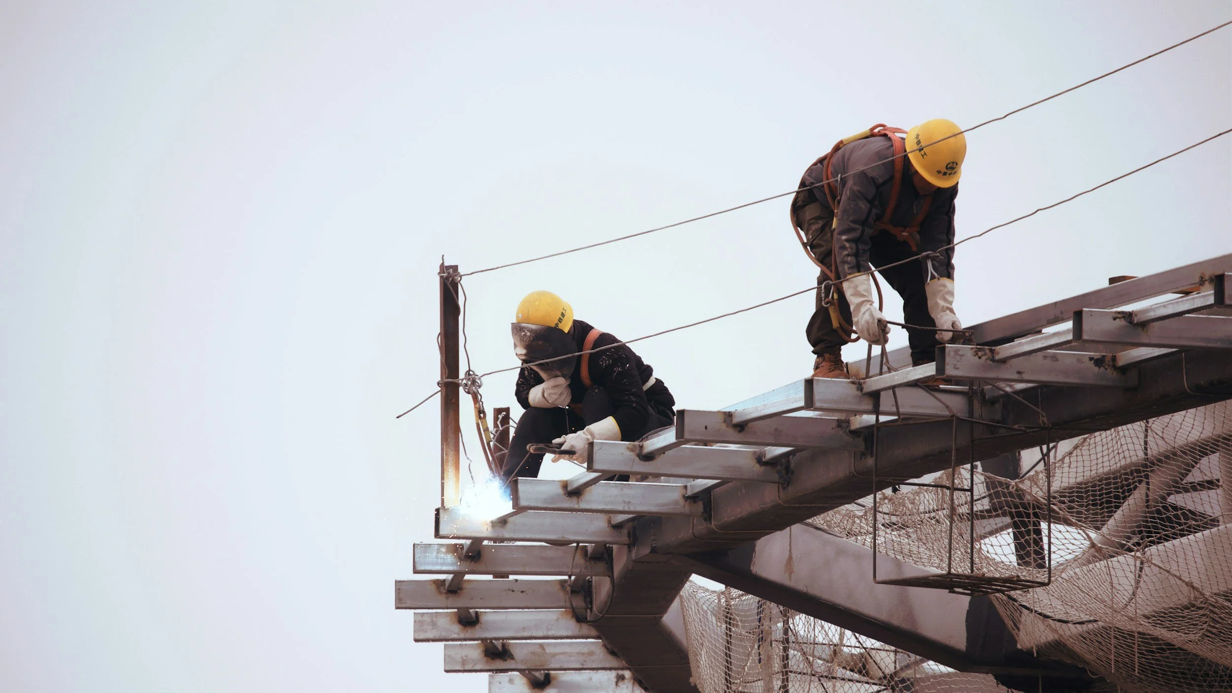 Two construction workers wearing yellow safety helmets and harnesses are working on a steel structure outside. One worker is welding while the other is handling tools.