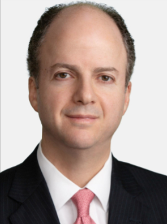 A professional man with short, dark, curly hair, wearing a dark suit, white shirt, and pink tie, against a light background.