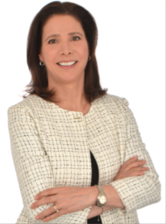 A smiling woman with shoulder-length dark hair, wearing a light-colored checkered blazer, standing with arms crossed against a white background.