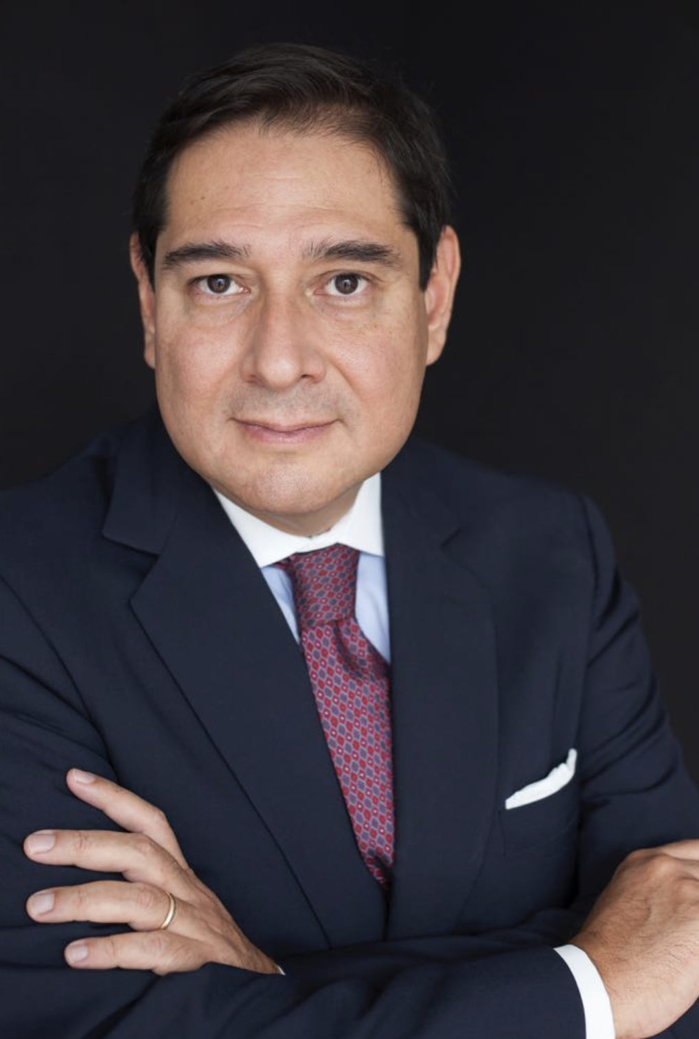 Headshot of a man in a dark suit and maroon tie, with crossed arms, against a black background.