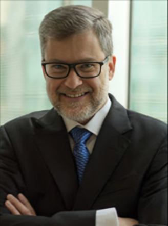 Professional man with gray hair and beard, wearing glasses, a dark suit, white shirt, and blue tie, smiling in an office setting.