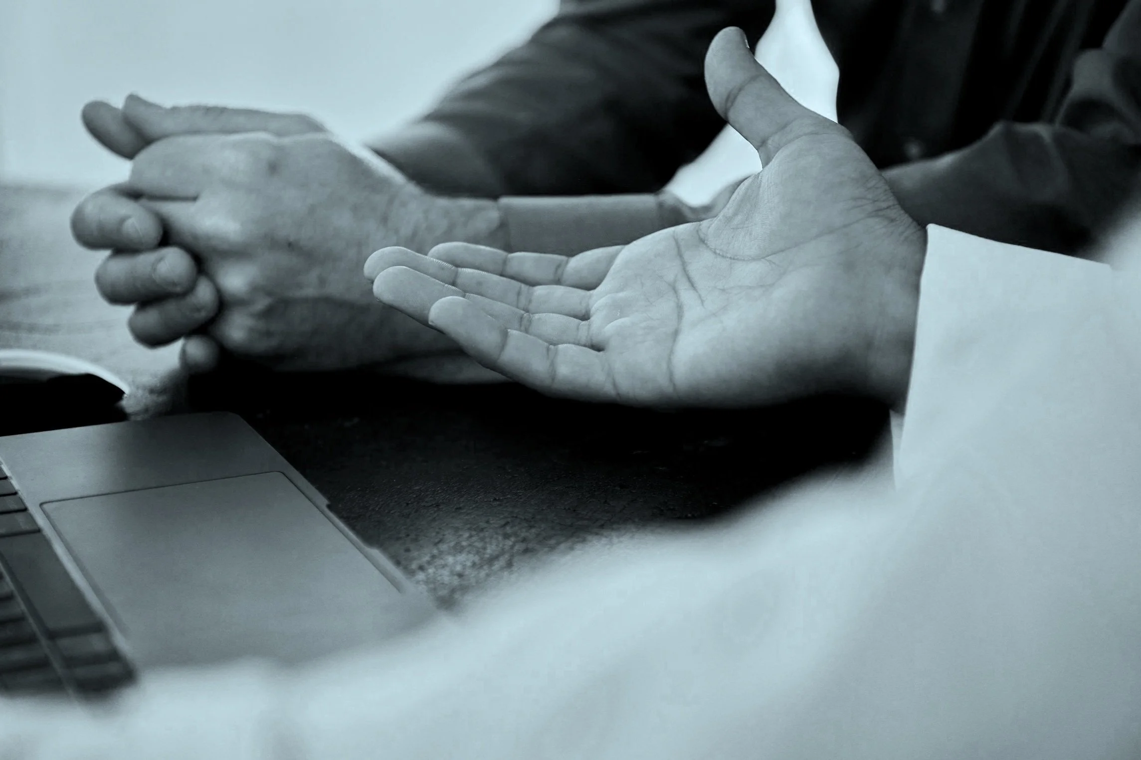 Two individuals with clasped hands are isolated from their faces, sitting at a table with a laptop in front. The image is in black and white.