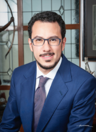 Professional man wearing glasses and a blue suit, sitting indoors with a background of large windows and decorative elements.