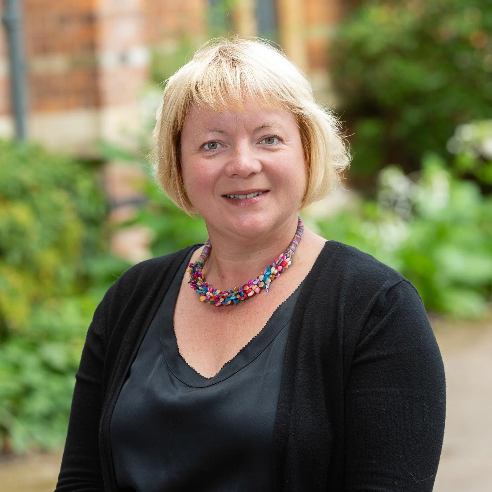 A middle-aged woman with blonde hair wearing a black blouse, colorful beaded necklace, and black cardigan, standing outdoors in front of green foliage and a brick building.