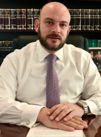 A man with a beard and shaved head dressed in a white shirt and purple tie sitting at a desk with a background of law books.