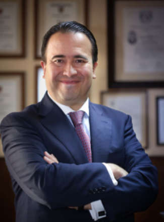 A man in a navy suit and patterned tie smiling with arms crossed, standing in front of framed certificates or awards.