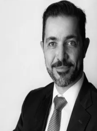 Black and white professional headshot of a man with dark hair and facial hair, wearing a suit and tie, smiling slightly.