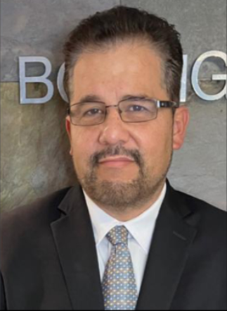 A man with glasses, short dark hair, and facial hair, wearing a dark suit and tie, standing in front of a gray wall with partial text visible.