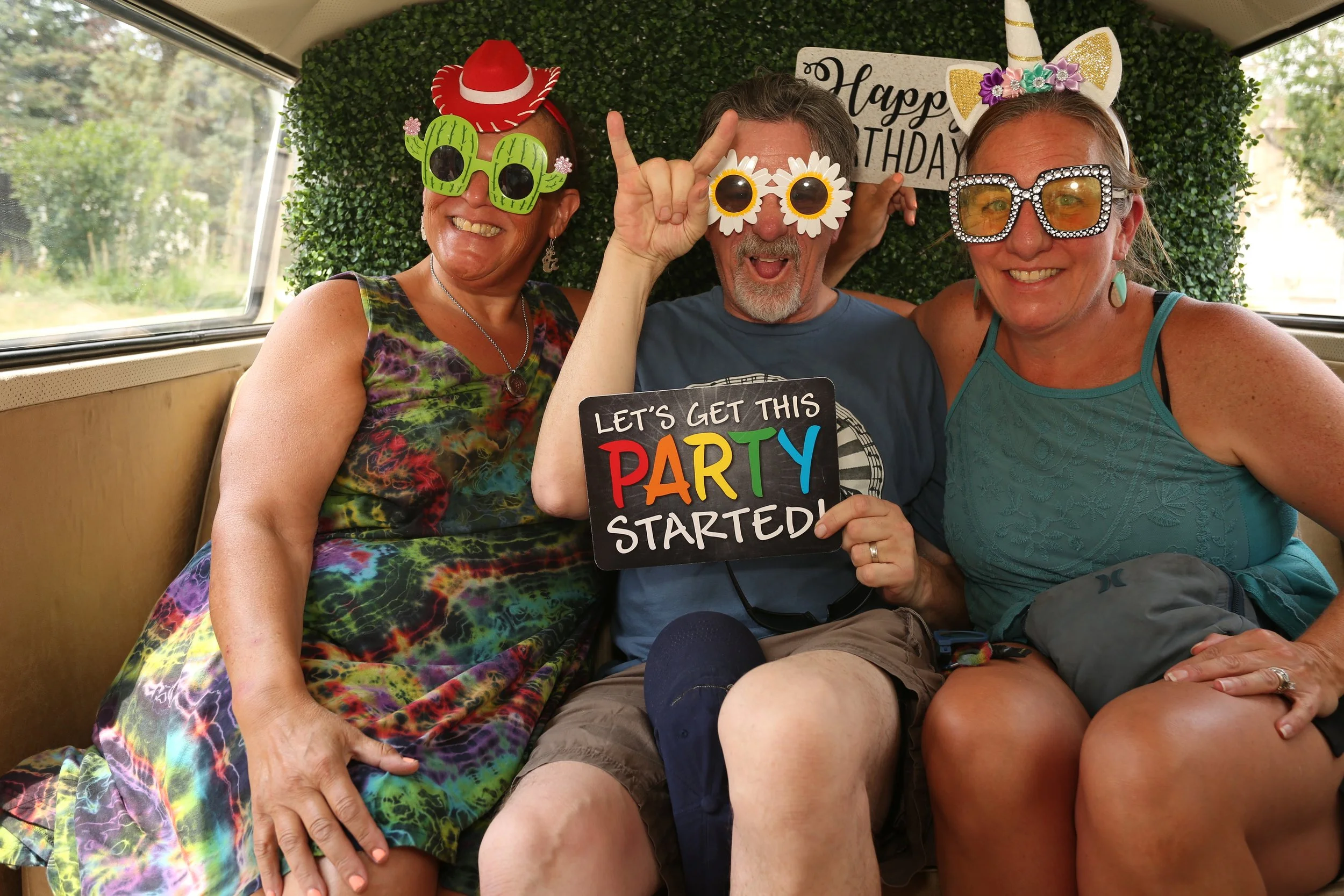 Three adults sitting together in a van, dressed festively for a birthday party with silly sunglasses and accessories. They are smiling, and one is holding a sign that reads "Let's get this party started." The background features a green wall with a "