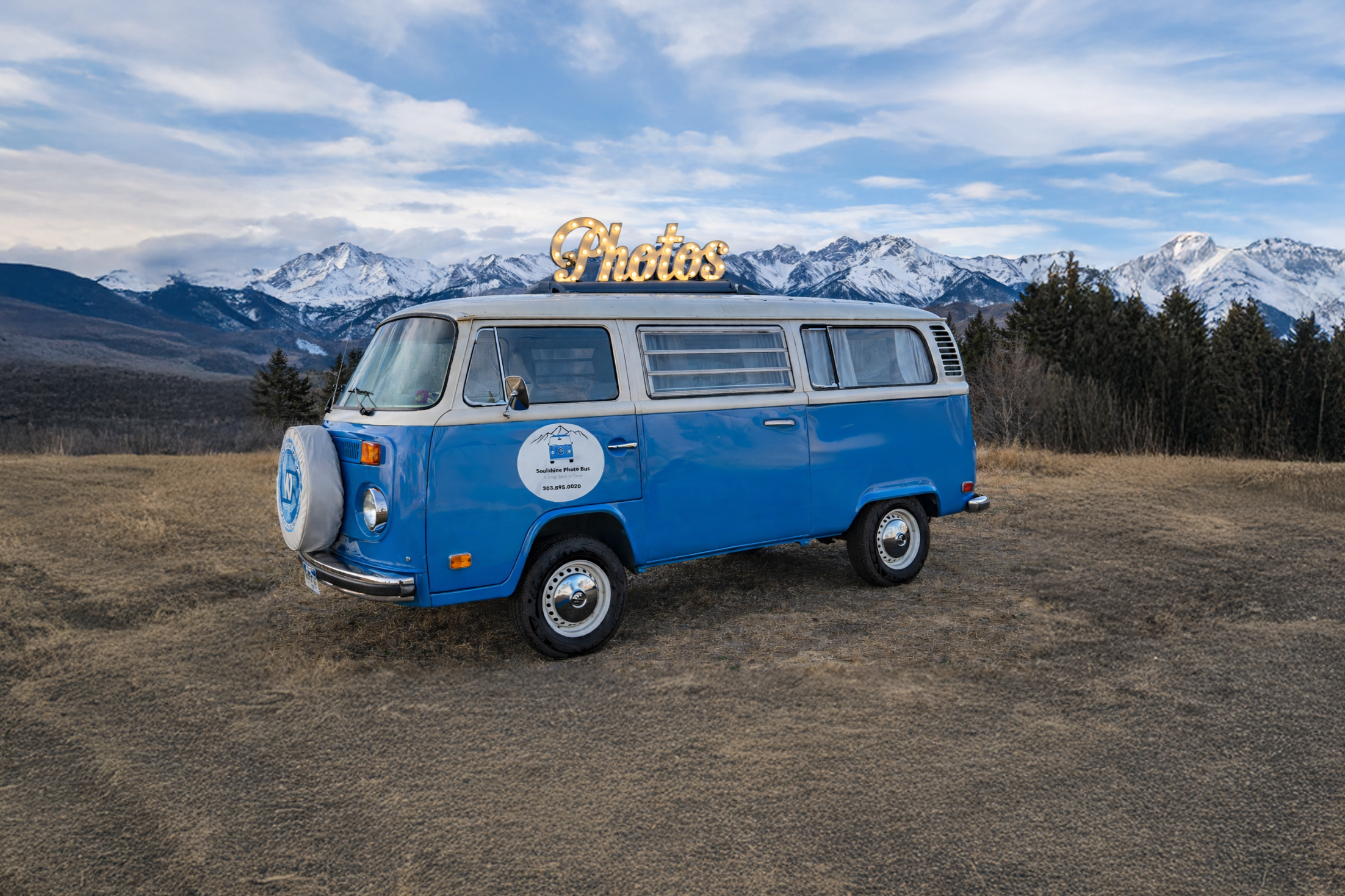 A vintage blue and white van with a illuminated 'Photos' sign on top, parked on a dirt field with mountains in the background.
