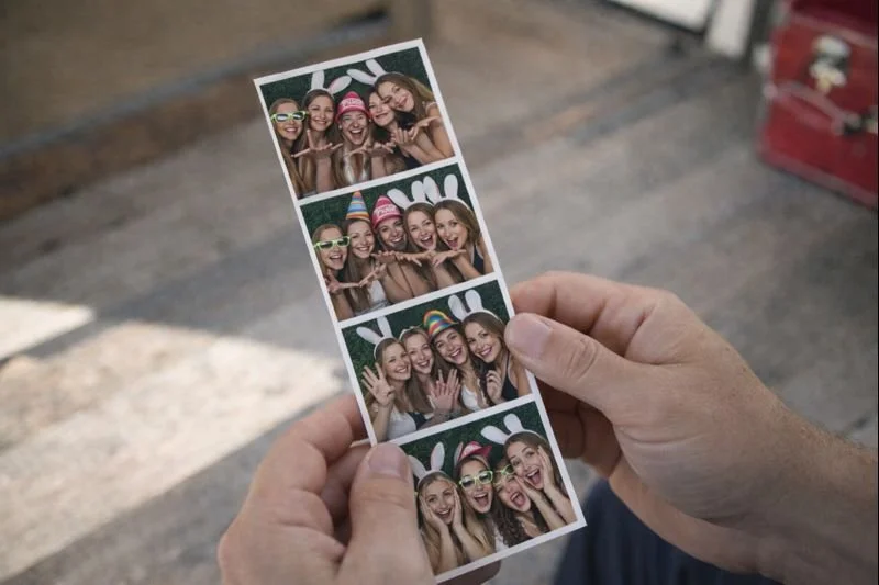 A person holding a photo strip with four pictures of four women wearing bunny ears and colorful glasses, making playful and happy expressions.