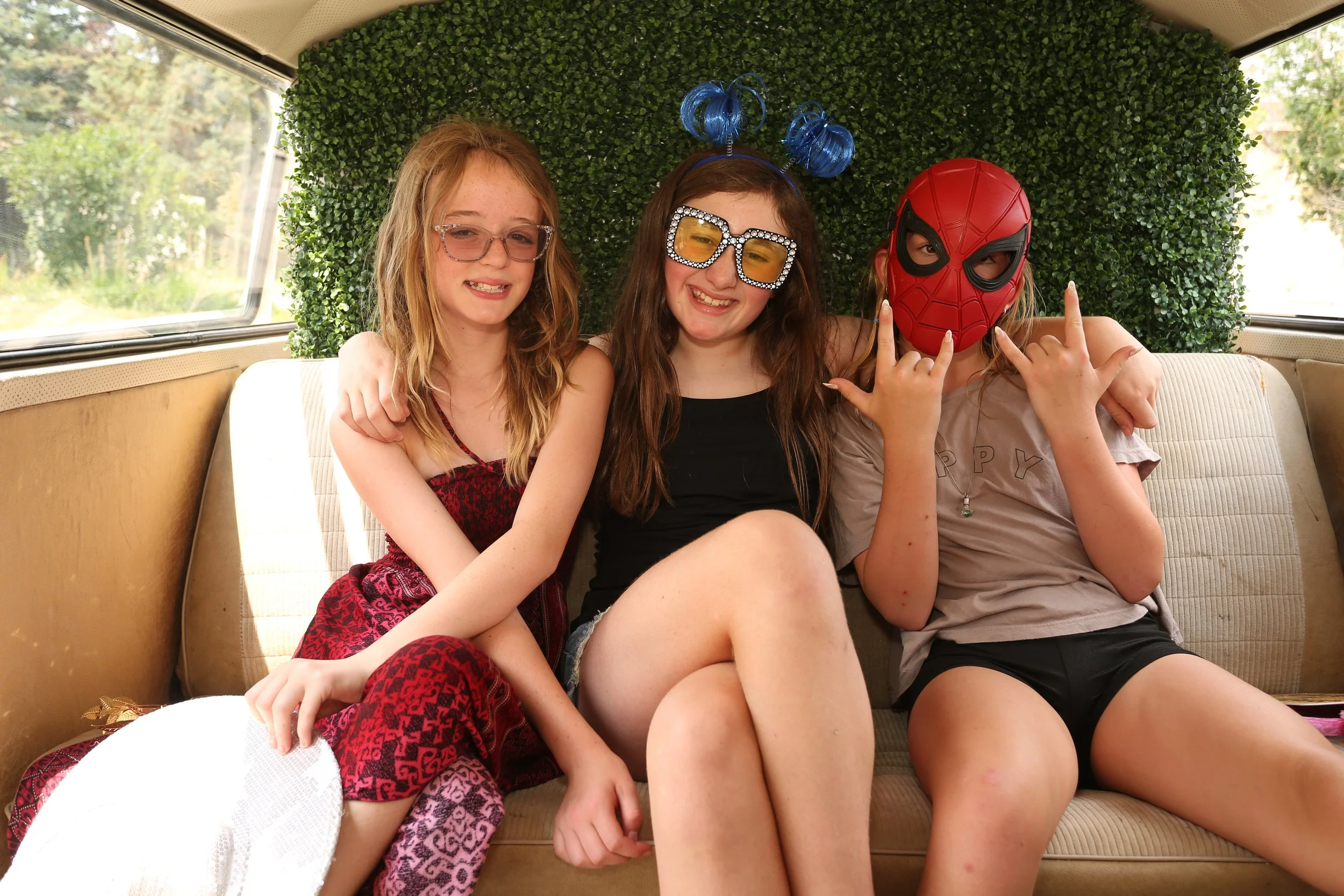Three young girls sitting on a vintage couch with a green foliage wall behind them. One girl is wearing a Spiderman mask and making rock and roll hand signs, while the other two girls are smiling, with one wearing large glasses with rhinestones and t