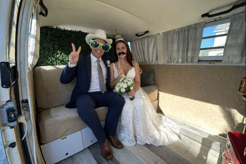 A couple dressed in wedding attire celebrating inside the photo booth bus, wearing fun accessories like oversized sunglasses, a party hat, and holding photo booth props.