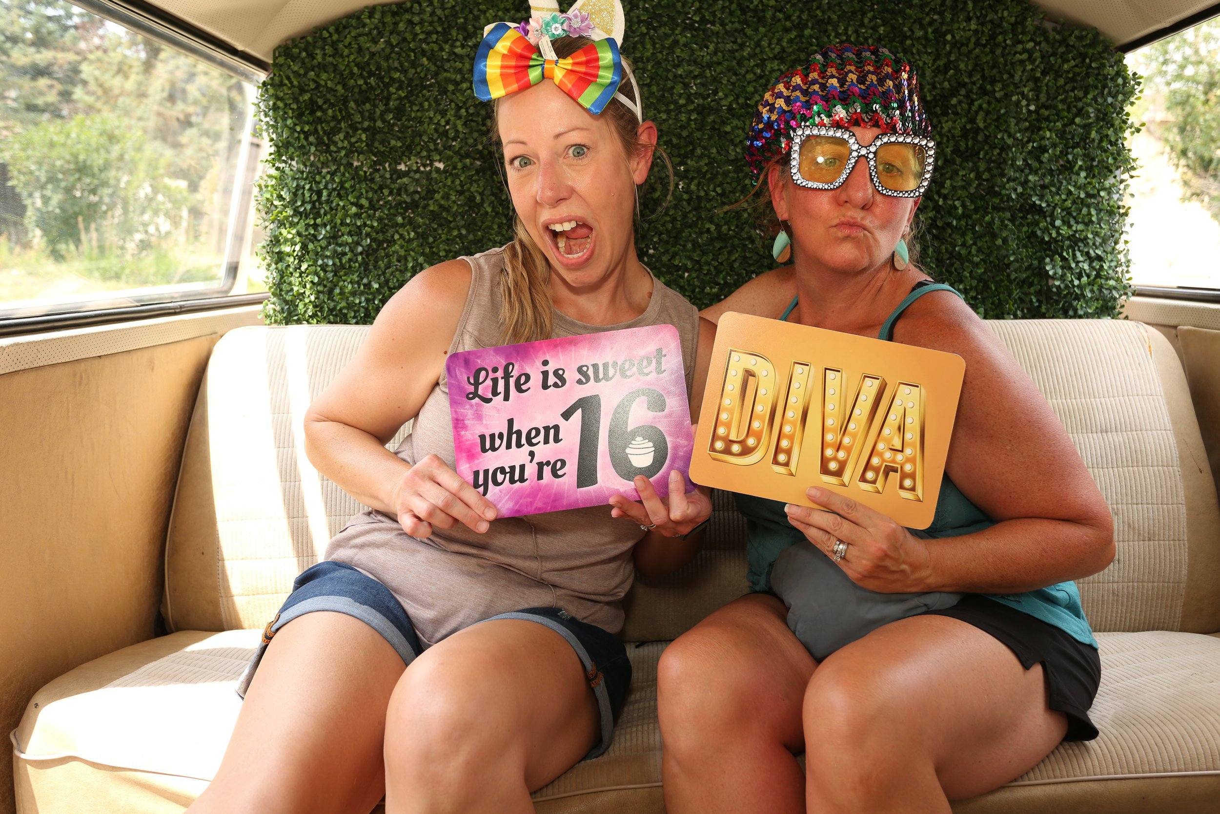 Two women sitting close together, holding signs and making playful expressions. The woman on the left has a large rainbow-colored bow headband and a sign that says 'Life is sweet when you're 16.' The woman on the right has a multicolored sequin hat, 