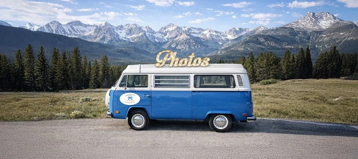 A vintage blue and white Volkswagen camper van with a large gold 'Photos' sign on top, parked on a mountain road with a picturesque landscape in the background.
