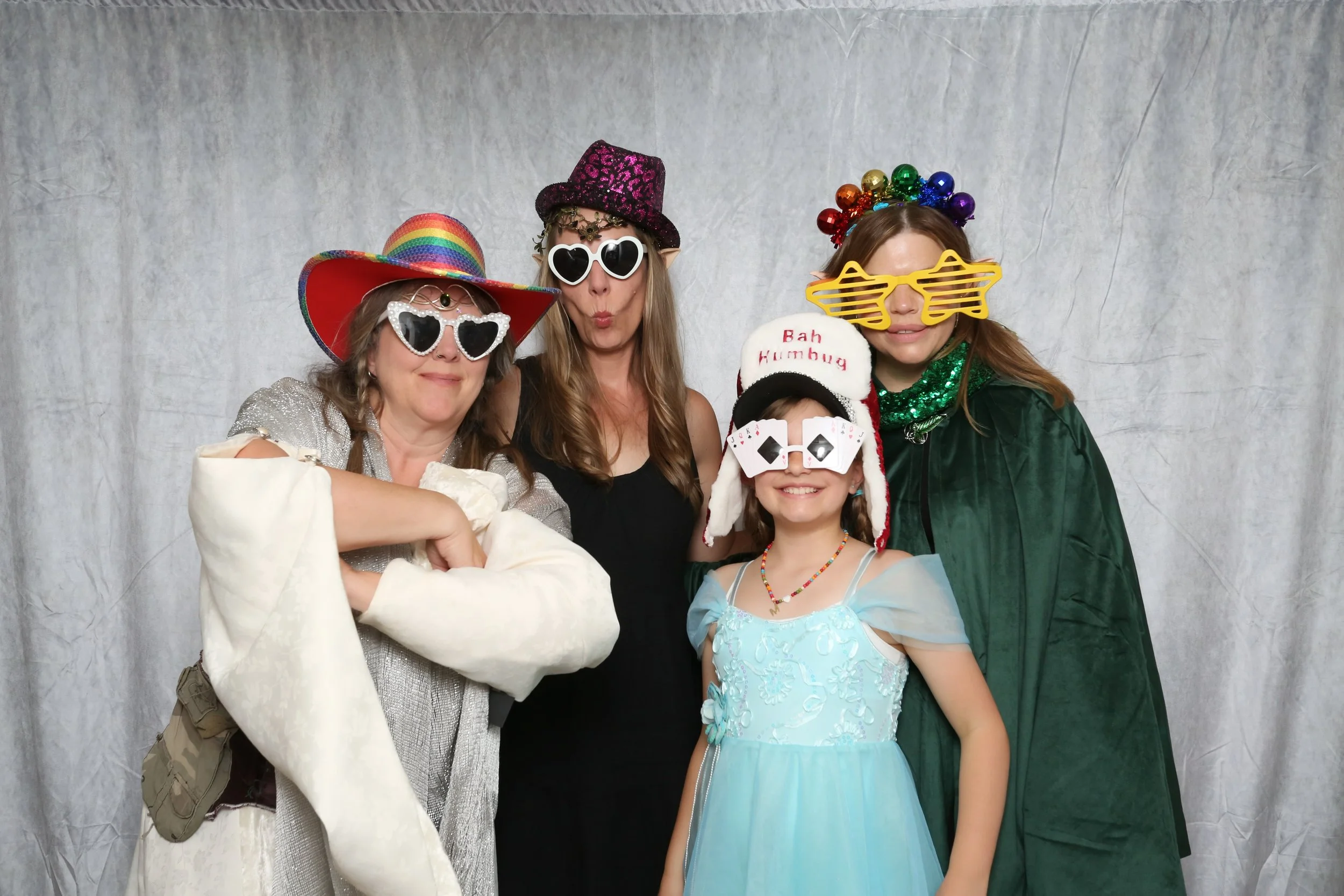 Group of five women and a young girl celebrating in a photo booth, wearing colorful costumes, hats, and sunglasses with playful accessories, smiling and making fun expressions.