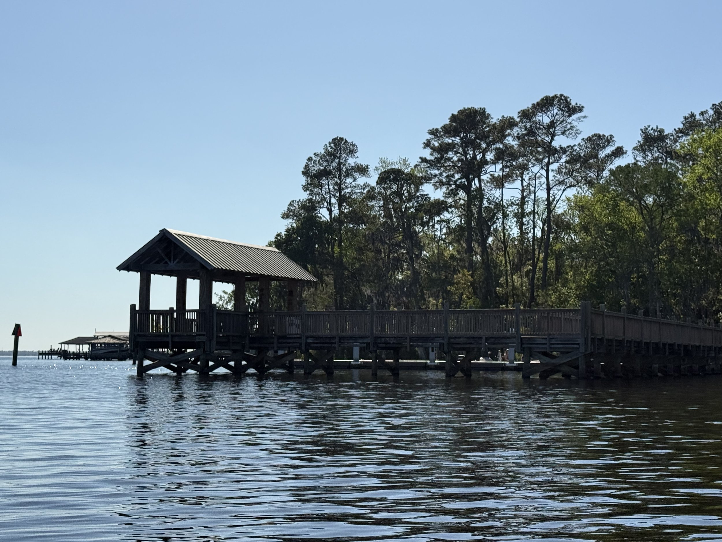 Wooden pier extending into a body of water with a small shelter at the end, surrounded by tall trees and a clear blue sky.