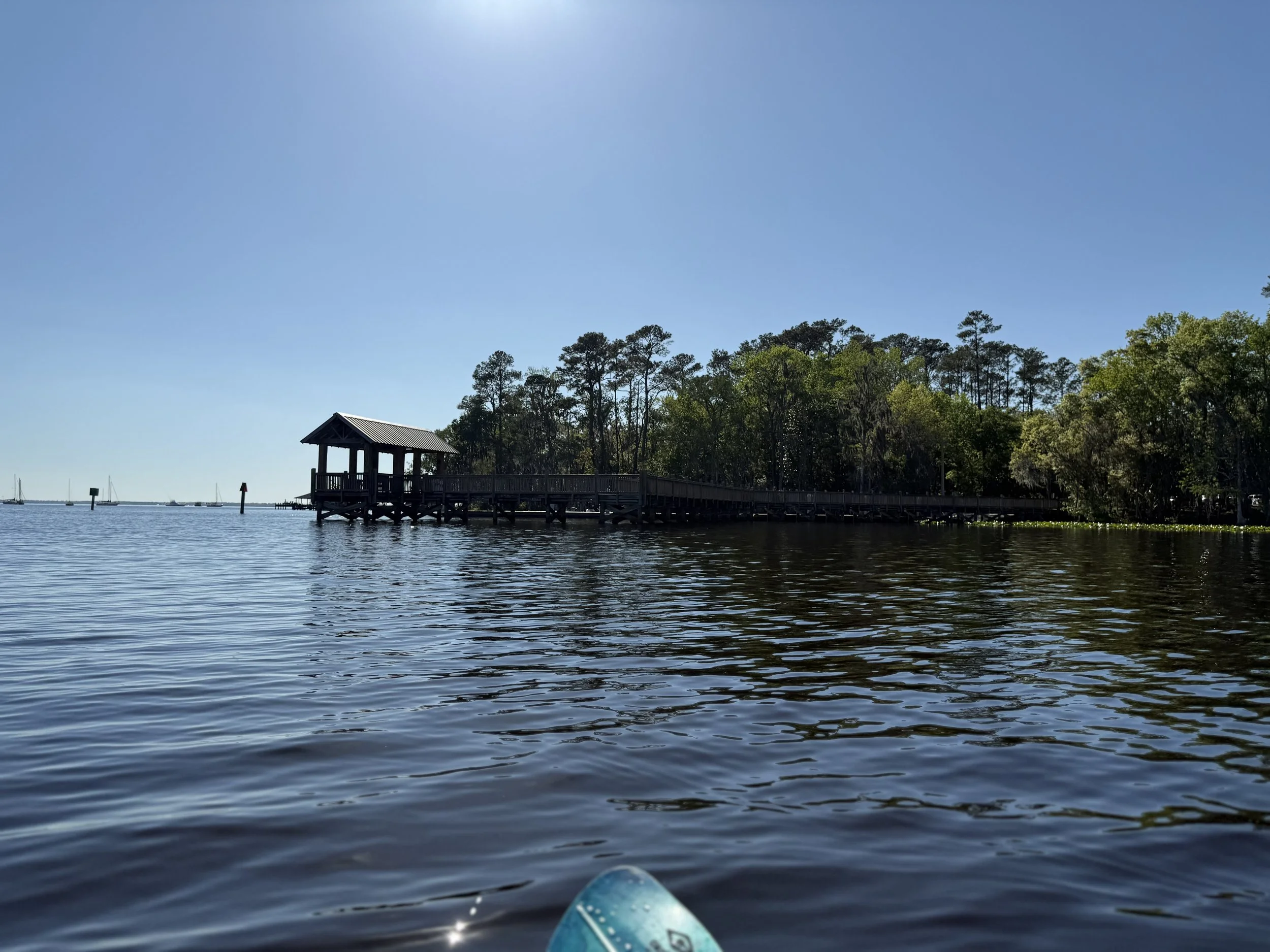 View of a wooden pier with a small covered shelter extending over calm water, with trees in the background under a clear blue sky.