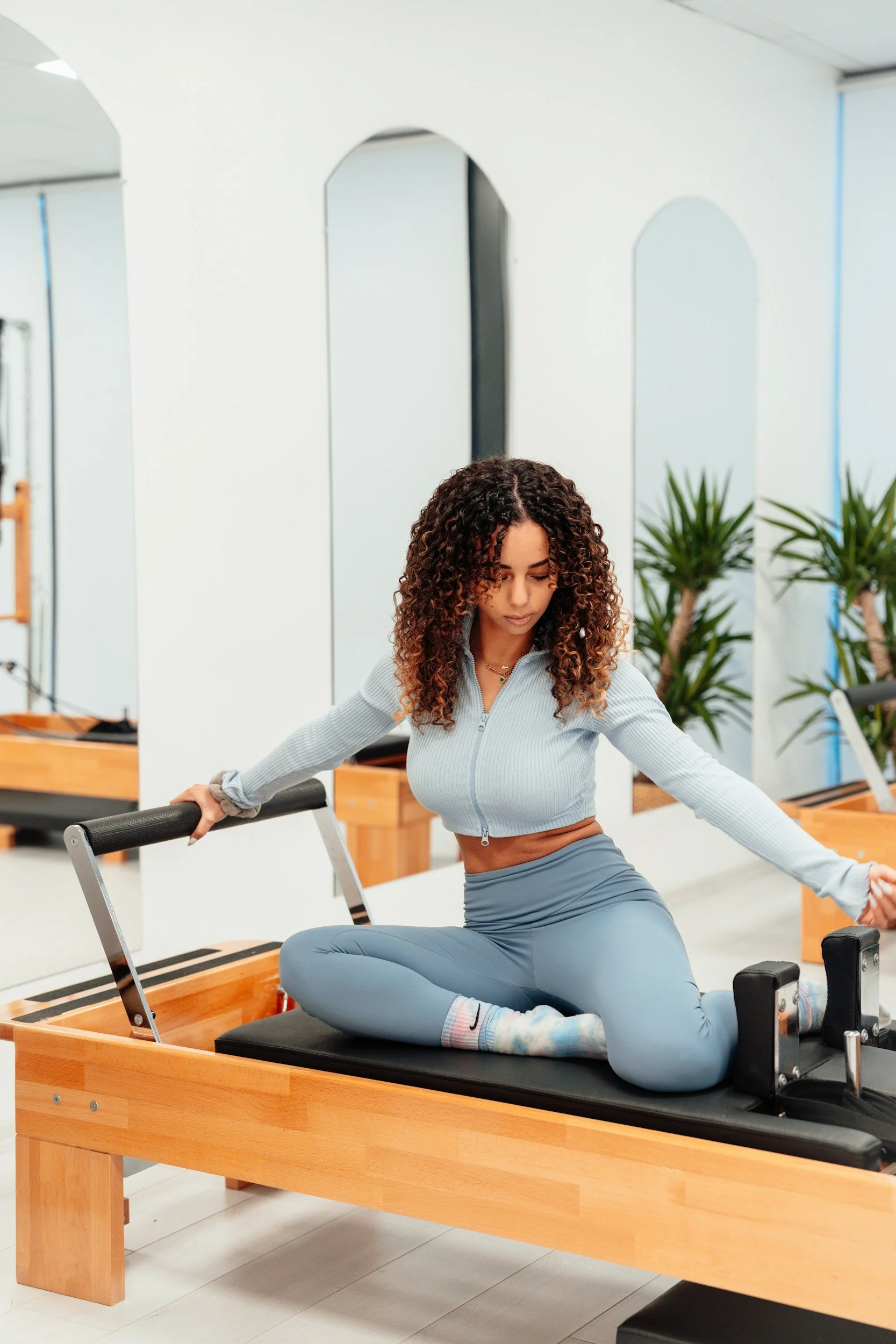 A woman with curly hair practicing Pilates on a wooden reformer machine in a bright, modern fitness studio with plants and mirrors.
