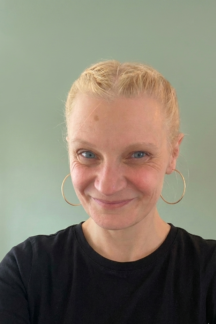 A woman with blonde hair in a braid, wearing large hoop earrings and a black shirt, smiling at the camera against a plain light green background.