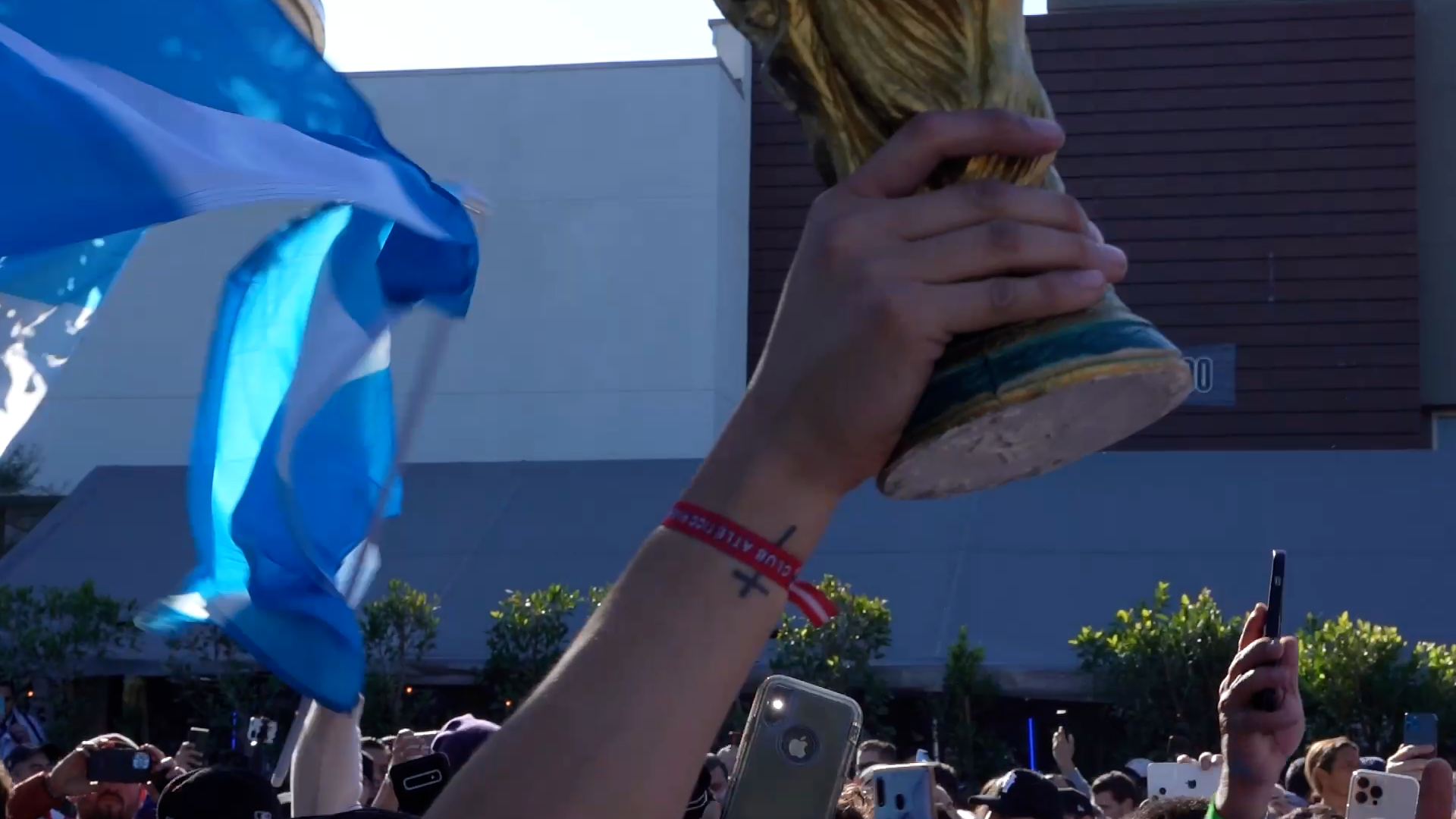 A crowd gathered outdoors during daytime, holding up their phones and a hand holding a ceramic trophy or statue with a wood-like appearance. One person in the crowd has a red wristband and a tattoo on their wrist. Others are capturing the scene with 