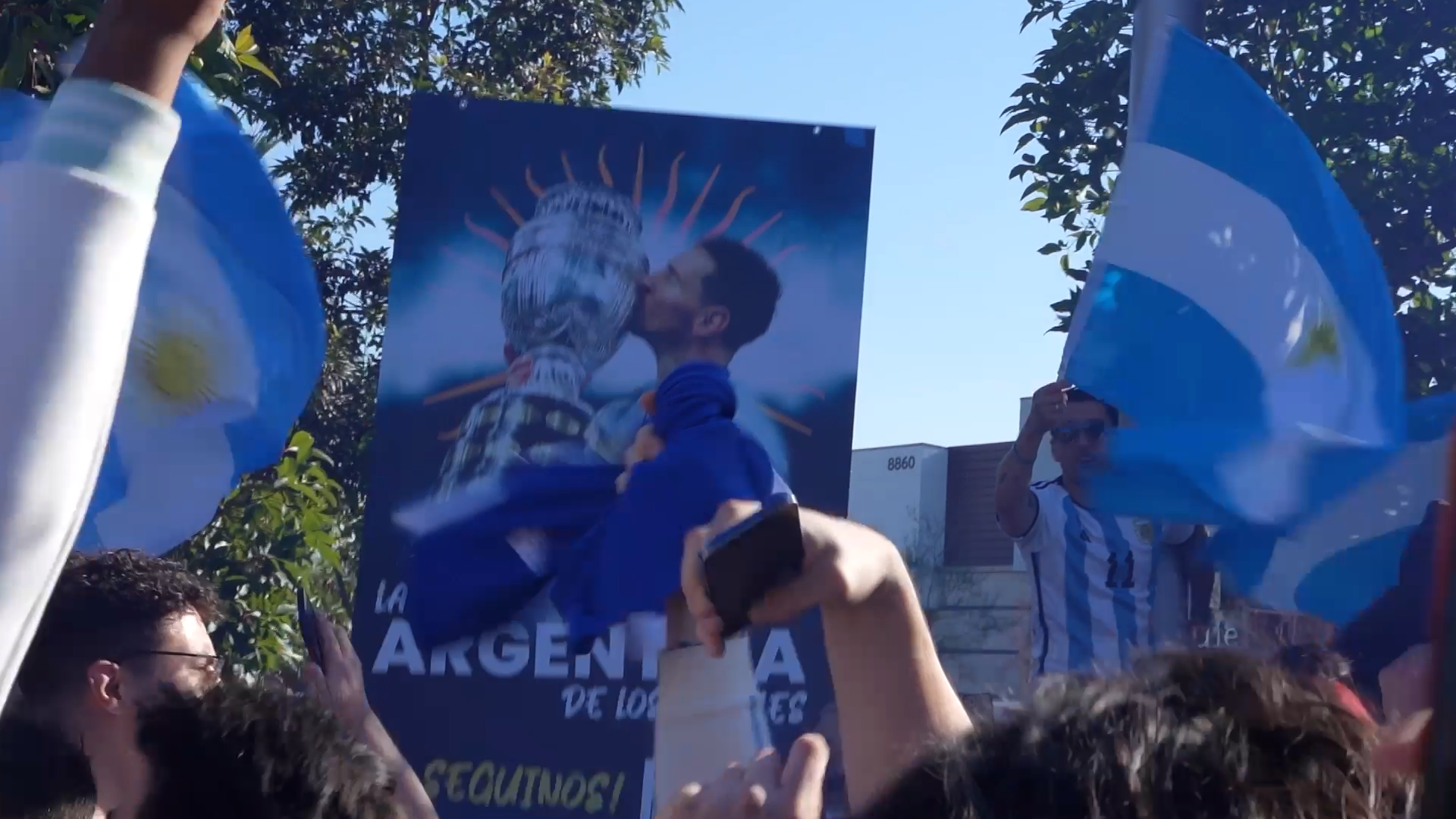 People holding flags and a poster of a soccer player, gathered outdoors during daytime, with trees and a building in the background.