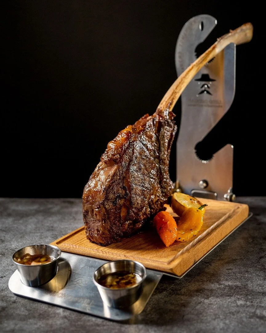 A cooked beef rib with a long bone, served on a wooden cutting board, with roasted vegetables and two small containers of dipping sauce, placed on a metal tray.