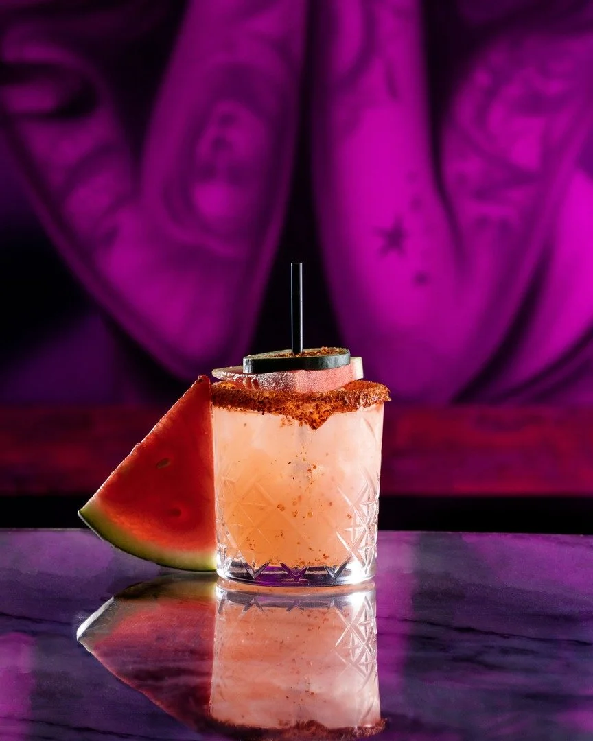 A watermelon-flavored cocktail in a glass with a salted rim, garnished with a watermelon wedge, on a reflective surface with a purple background.