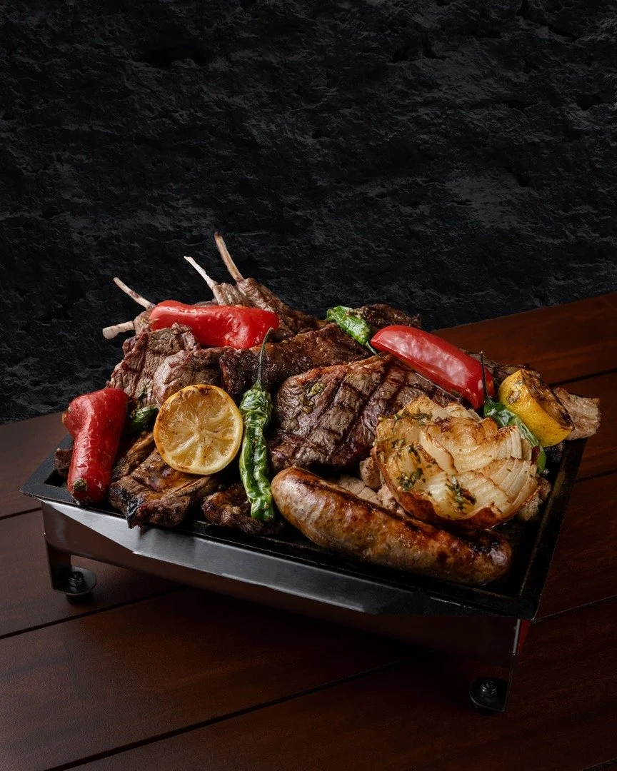 A black tray with cooked meats, grilled vegetables including red and green peppers, a lemon wedge, and grilled onion on a wooden table against a dark background.