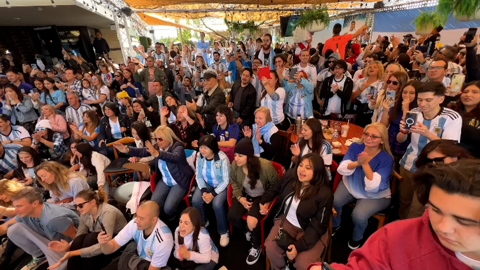 A large crowd of people gathered indoors, many wearing Argentina soccer jerseys, some taking photos and videos, some clapping and smiling, in a festive environment with decorations and hanging plants.
