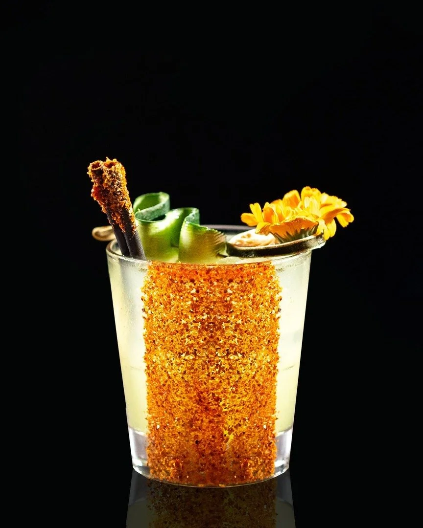 A cocktail glass featuring a yellow drink, garnished with a lemon twist, cucumber ribbons, a breaded and fried skewer, and a yellow flower, set against a black background.