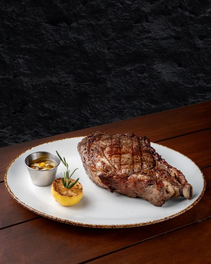 A large grilled steak on a white plate, served with a small metal cup of sauce and a grilled lemon half garnished with a sprig of rosemary.