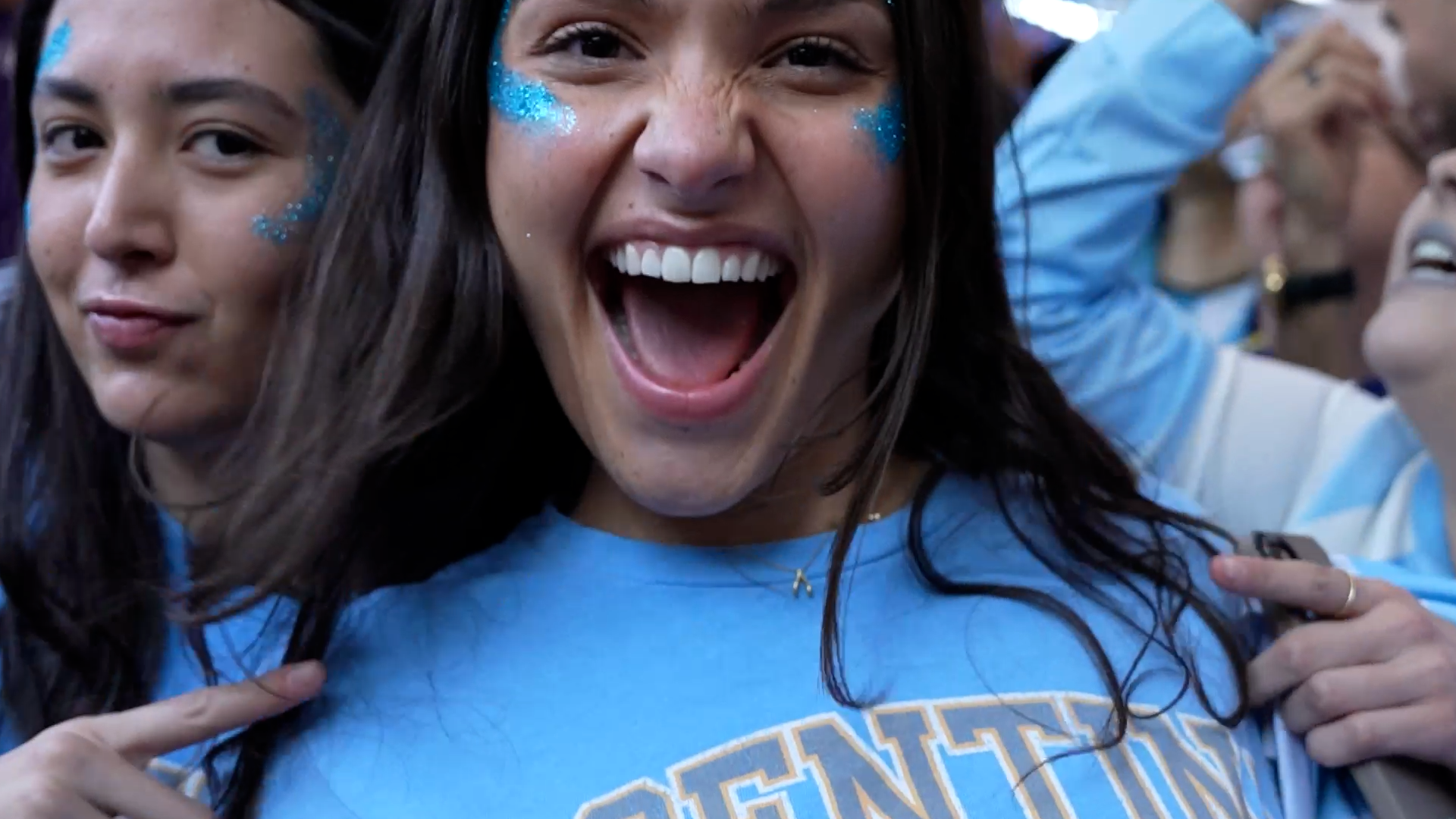Group of women at a gathering, one woman in the center showing excitement and joy with glitter face paint, holding a phone, wearing a blue T-shirt, surrounded by others with similar attire.