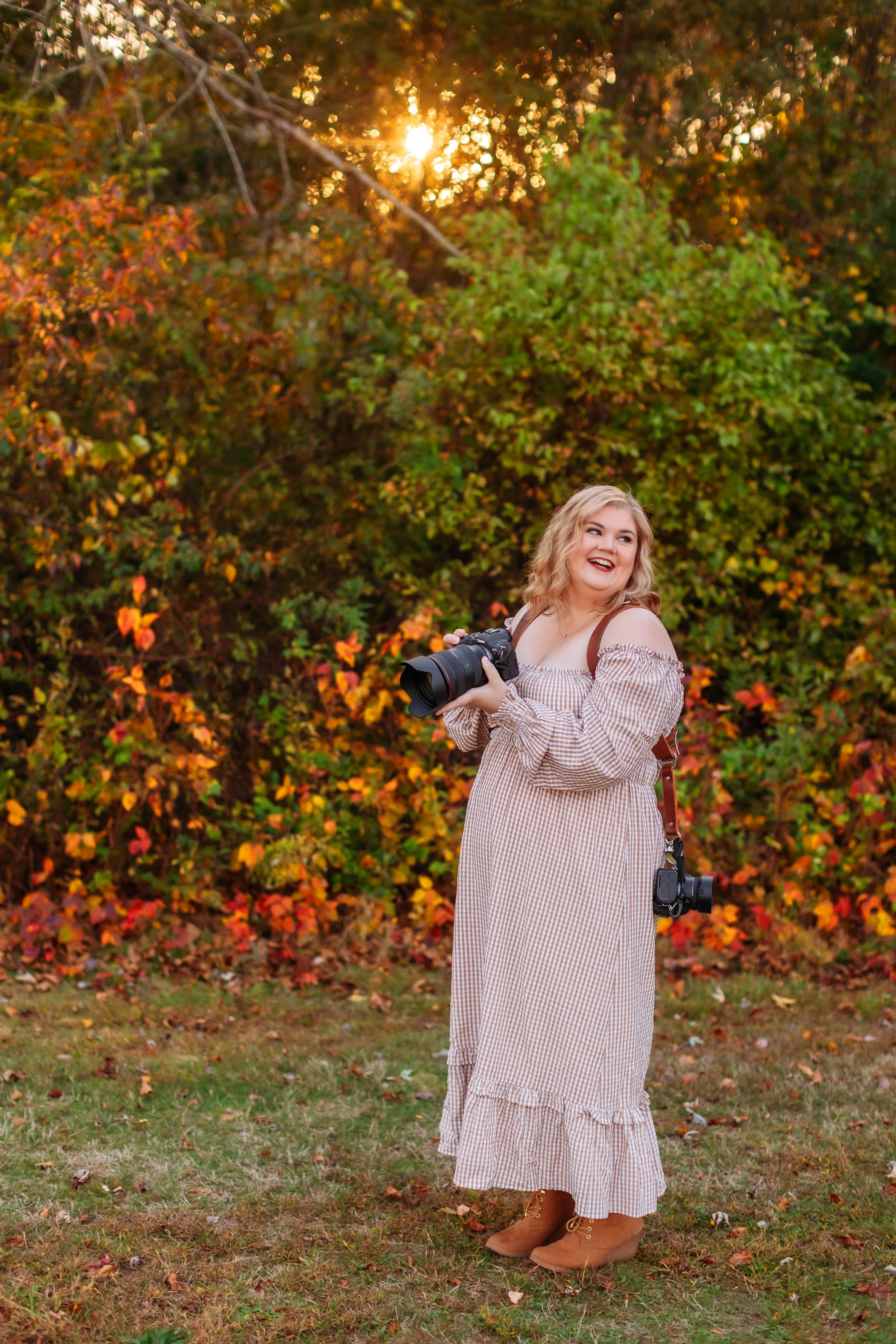A woman with blonde curly hair in a long brown and cream gingham dress and brown boots, holding a camera with a large lens, standing outside in front of fall foliage during sunset, smiling.