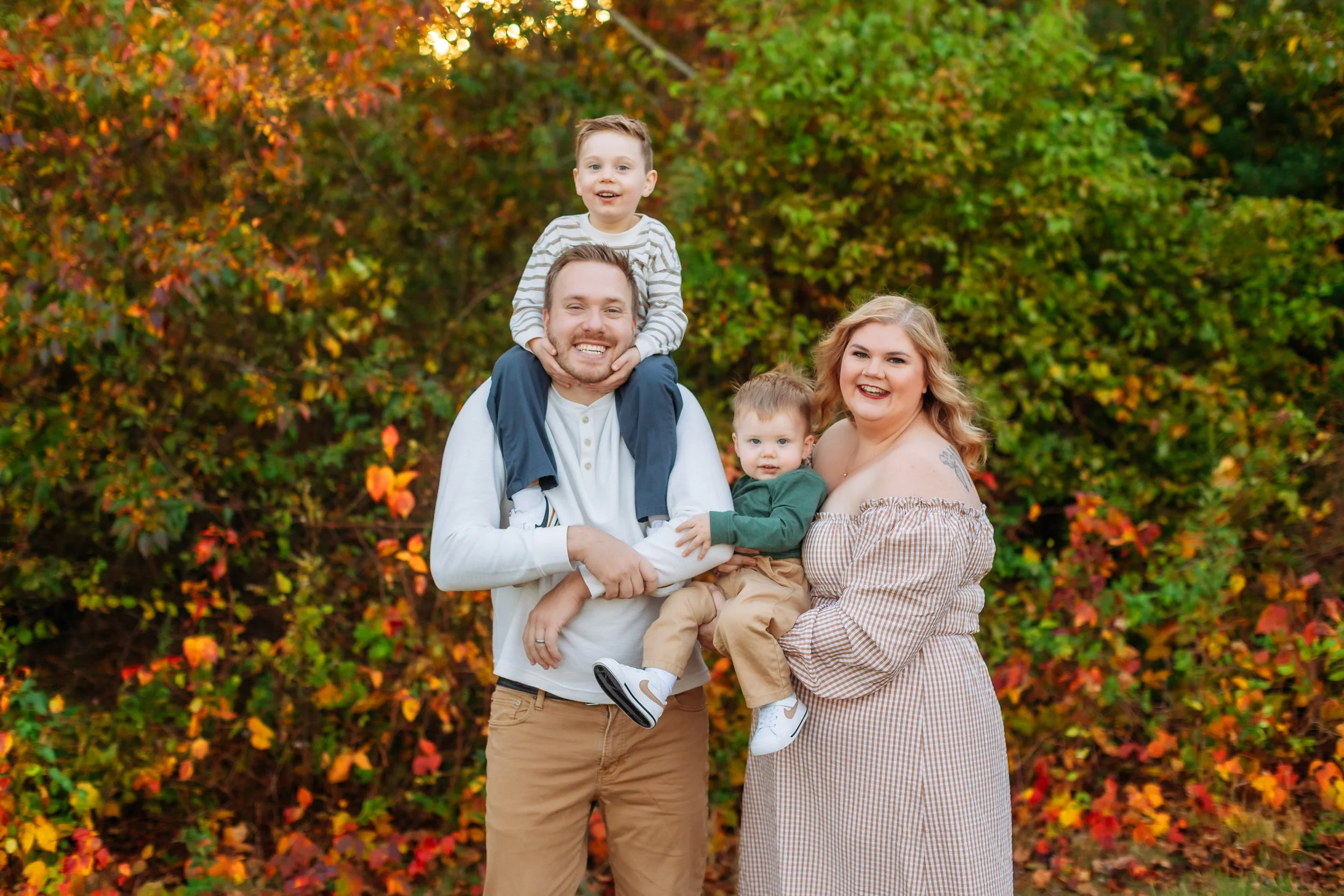 A family of four smiling outdoors in front of autumn foliage. The father is holding one child on his shoulders, and the mother is holding the other child. All are smiling and dressed casually for fall.