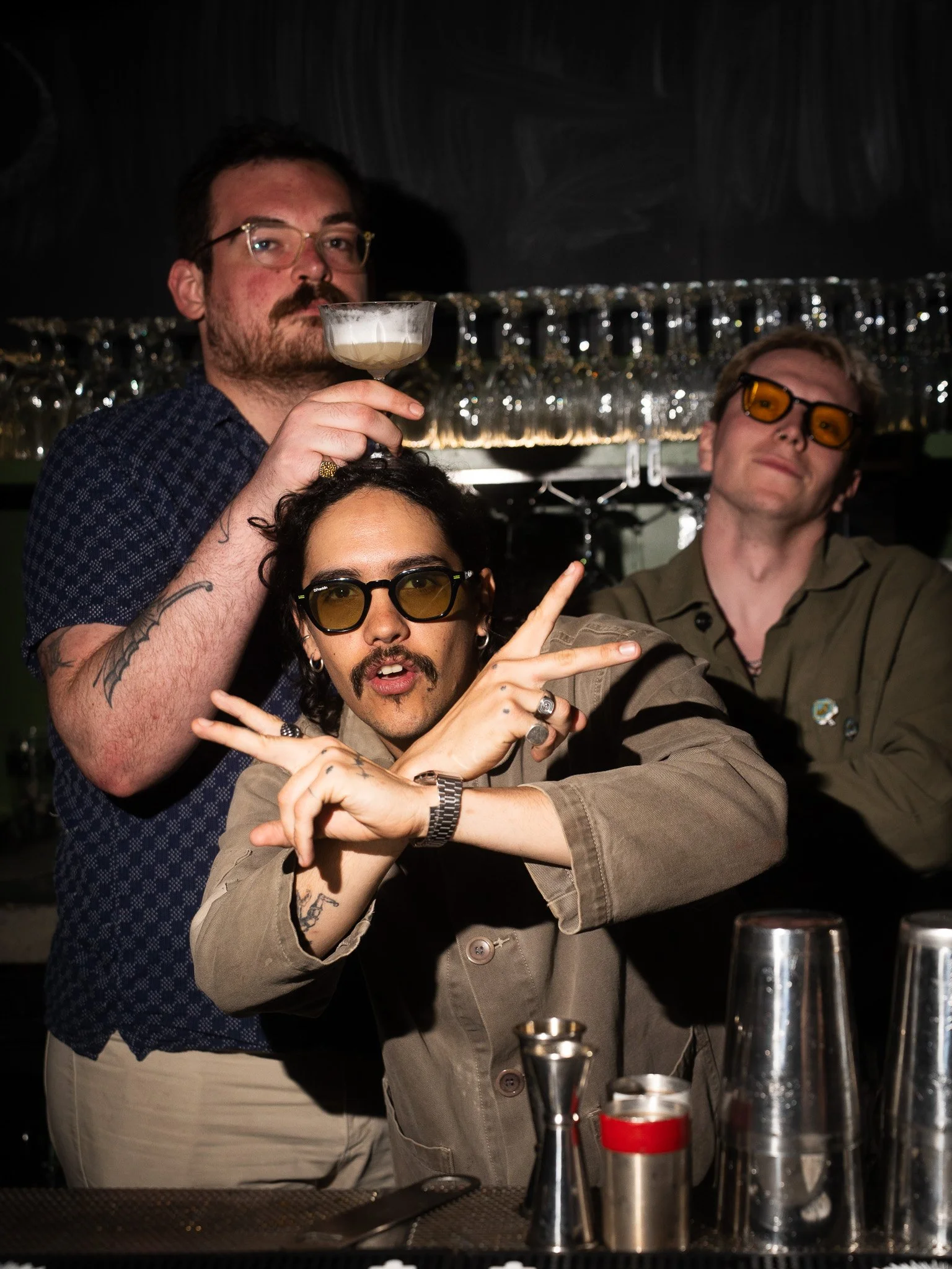 Three young men posing at a bar, one holding a drink and the other two making peace signs, all wearing sunglasses.