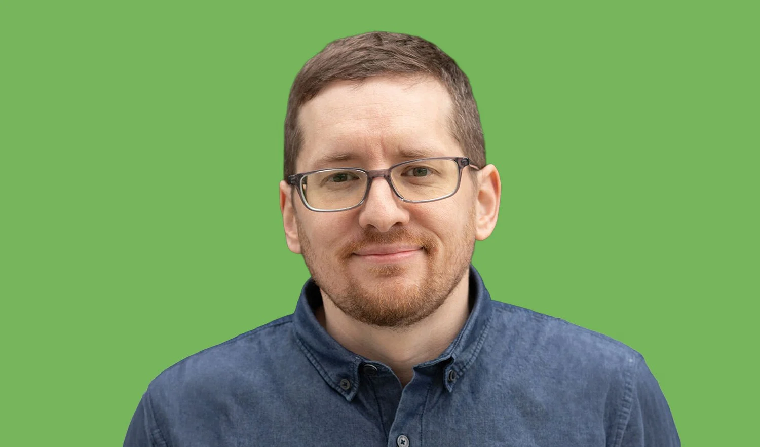 A man with glasses and a beard smiling, wearing a denim shirt, with a green background.